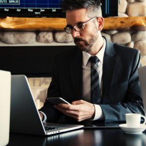 man in suit in front of a laptop searching for executive resumes online while holding a smartphone